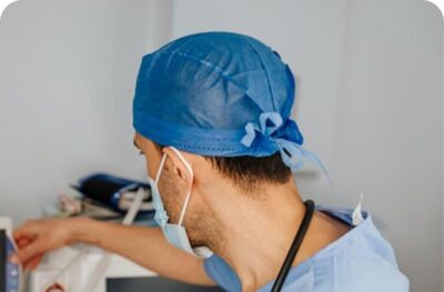 Surgeon in a blue scrub cap and mask, seen from behind, reaching towards equipment in an operating room